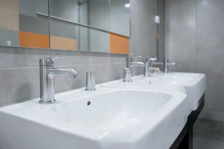 Ambiente de banheiro público com várias torneiras antivandalismo instaladas em pias de cerâmica.